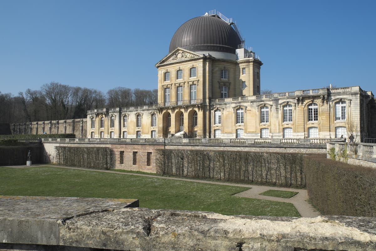 Grand bâtiment classique en pierre surmonté d'une coupole sombre, devant une pelouse et des haies taillées.