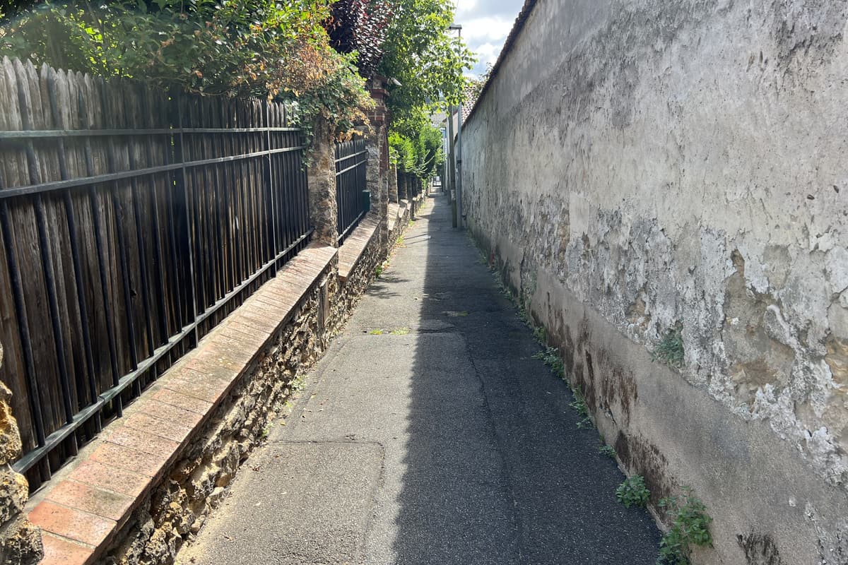 Sente étroite entre un muret de meulière surmonté d'une grille et un haut mur ancien enduit.