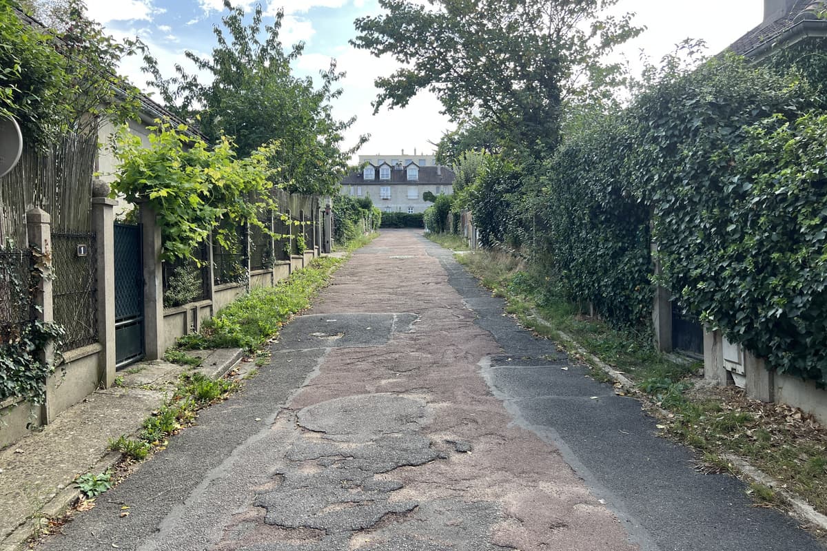 Voie privée étroite entre des clôtures, des haies et des grilles, menant à une maison à lucarnes.