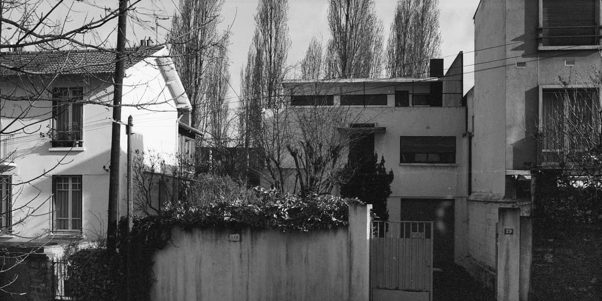 Photographie en noir et blanc : maison blanche aux volumes cubiques et bandeaux de fenêtres, derrière un mur et un portail, entre deux maisons voisines.